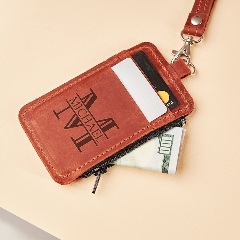 Puede incluir: Una cartera de cuero marrón con un bolsillo con cremallera y una ranura para tarjetas. La cartera tiene una inscripción personalizada que dice 'MICHAEL T.V.I.'