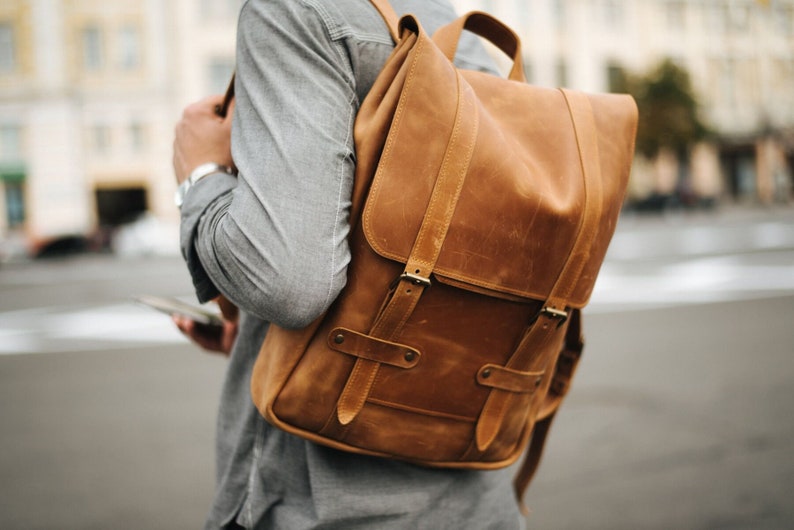 May include: A brown leather backpack with two adjustable straps and buckles. The backpack has a flap with a buckle closure.
