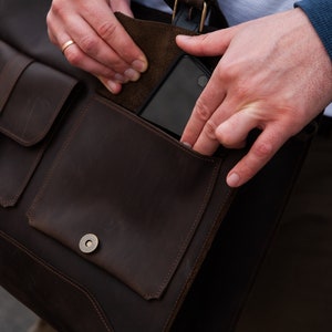 May include: A brown leather messenger bag with a flap closure and a buckle. The bag has a large pocket on the front and a smaller pocket on the side. A person is reaching into the side pocket.