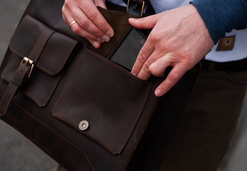 May include: A brown leather messenger bag with a flap closure and a buckle. The bag has a large pocket on the front and a smaller pocket on the side. A person is reaching into the side pocket.