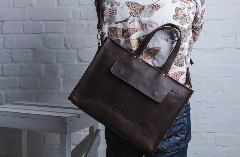 K&ouml;nnte beinhalten: Eine braune Ledertasche mit einer Fronttasche und einem langen Riemen. Die Tasche wird &uuml;ber der Schulter einer Person getragen, die ein wei&szlig;es Hemd mit einem Schmetterlingsmuster und eine blaue Jeans tr&auml;gt.