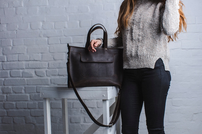 K&ouml;nnte beinhalten: Eine braune Ledertasche mit einem langen Trageriemen. Die Tasche hat eine Fronttasche und steht auf einem wei&szlig;en Tisch.