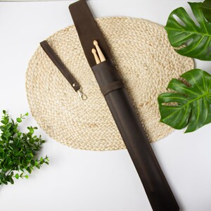May include: A brown leather drumstick case with a strap and two wooden drumsticks inside. The case is lying on a woven mat.