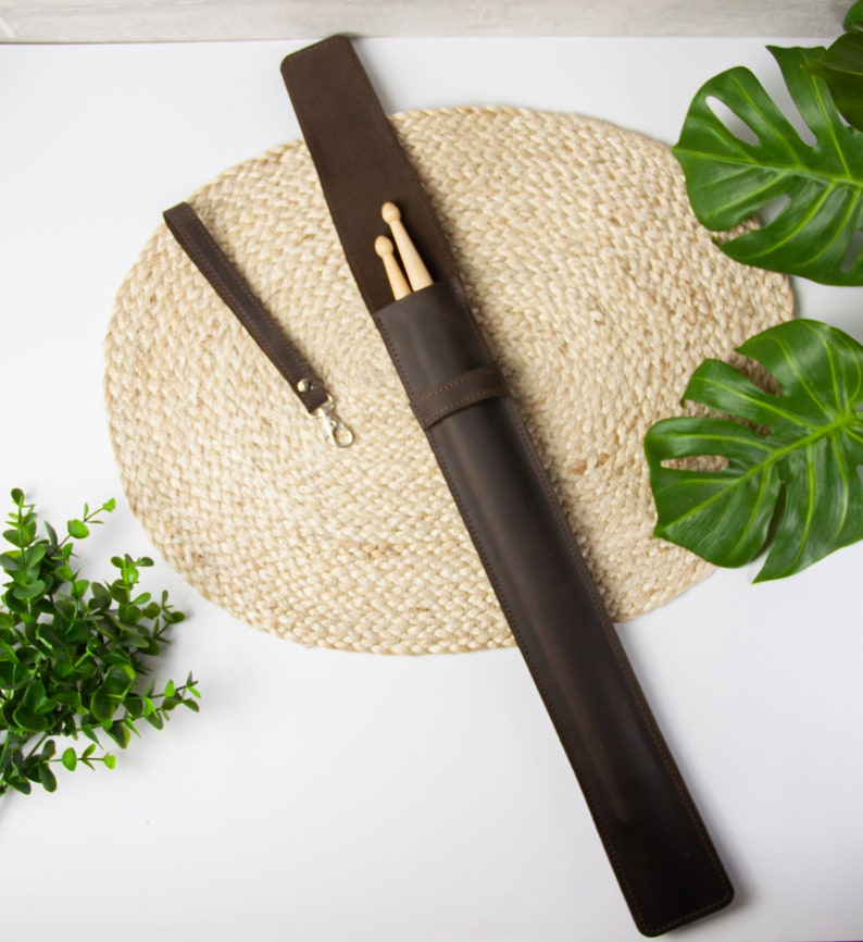 May include: A brown leather drumstick case with a strap and two wooden drumsticks inside. The case is lying on a woven mat.