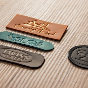 Può includere: Quattro etichette in pelle con diversi design e testi. L'etichetta superiore è marrone con testo dorato che recita "A road by the Lord". La seconda etichetta è blu turchese con un design a foglia verde e il testo "SLG". La terza etichetta è nera con il testo "TWISS REALTY". La quarta etichetta è nera con un design a farfalla stilizzato e il testo "EB".