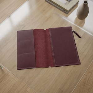 Può includere: Una copertina per taccuino in pelle color bordeaux scuro con finitura esterna liscia, interno aperto con molteplici fessure per carte sul lato sinistro, una tasca a tutta lunghezza a destra e un cursore della cerniera in metallo argentato.