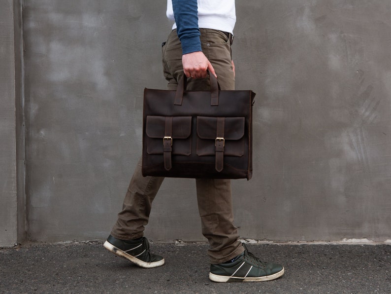 May include: A brown leather briefcase with two front pockets and a top handle. The briefcase is being carried by a person wearing brown pants and a blue shirt.