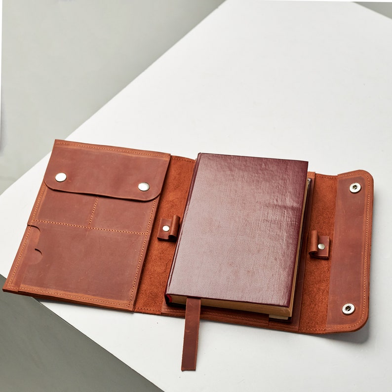 K&ouml;nnte beinhalten: Eine braune Leder-Notizbuchh&uuml;lle mit mehreren Taschen und einem burgunderfarbenen Notizbuch im Inneren. Die H&uuml;lle hat silberne Druckkn&ouml;pfe und einen Lederriemen.