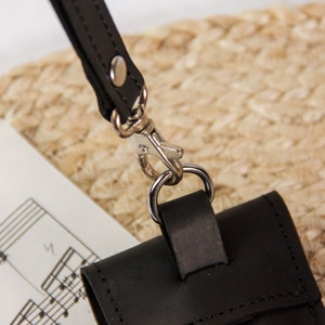 May include: A black leather phone case with a silver metal clasp and a black leather strap. The case is lying on a piece of sheet music.