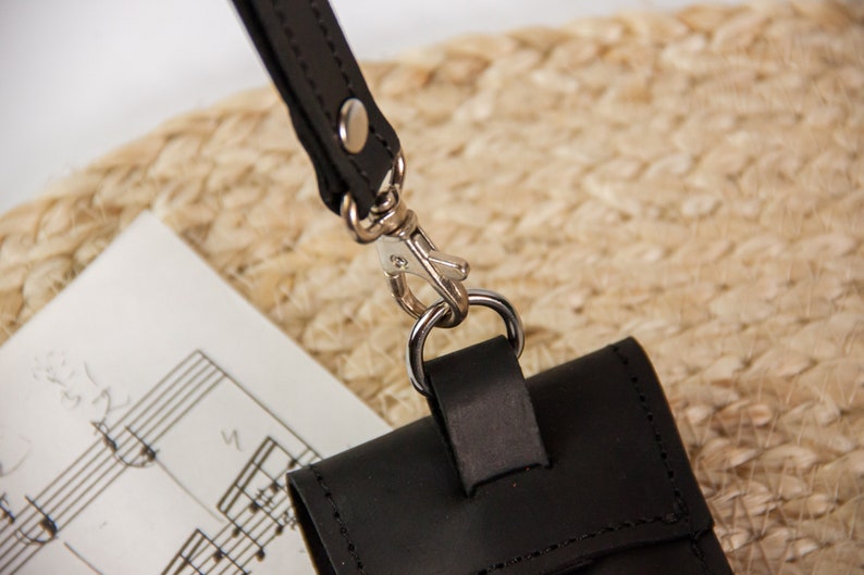 May include: A black leather phone case with a silver metal clasp and a black leather strap. The case is lying on a piece of sheet music.