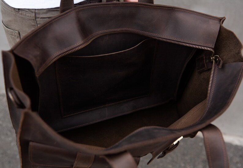 May include: A brown leather tote bag with a zippered top and a smaller pocket inside. The bag has two straps and a buckle on the front.