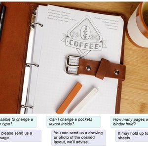 May include: A brown leather binder with a metal ring closure and a leather strap. The binder has a white page with a drawing of a coffee cup and the text "1920 COFFEE". The binder is on a wooden surface with a cup of tea and two colored pencils.