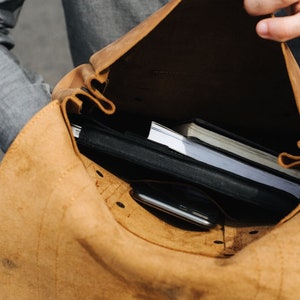 May include: A brown leather backpack with a flap open revealing a black notebook, a white notebook, and a black smartphone inside.