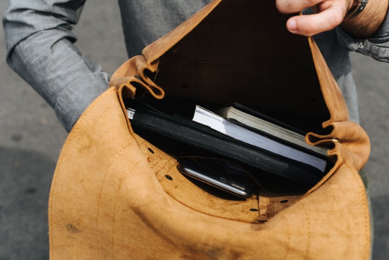 May include: A brown leather backpack with a flap open revealing a black notebook, a white notebook, and a black smartphone inside.
