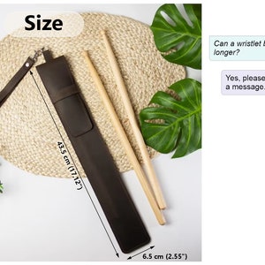 May include: Brown leather drumstick holder with a wrist strap. The holder measures 43.5 cm long and 6.5 cm wide. The text "Size" is written in white on a black background. The text "Can a wristlet be longer?" and "Yes, please send us a message." are written in white on a light blue background.
