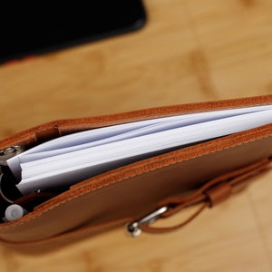 May include: A brown leather bound journal with a white paper insert and a silver ring binder. The journal is closed and has a strap closure.