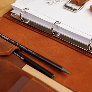 May include: A brown leather binder with a silver ring mechanism. The binder is open and has a few sheets of white paper inside. There are two pencils and a small orange eraser on the cover.