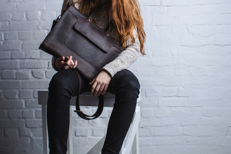 K&ouml;nnte beinhalten: Eine braune Ledertasche mit Klappenverschluss und einer Fronttasche. Die Tasche wird von einer Person getragen, die schwarze Hosen und einen wei&szlig;en Pullover tr&auml;gt.