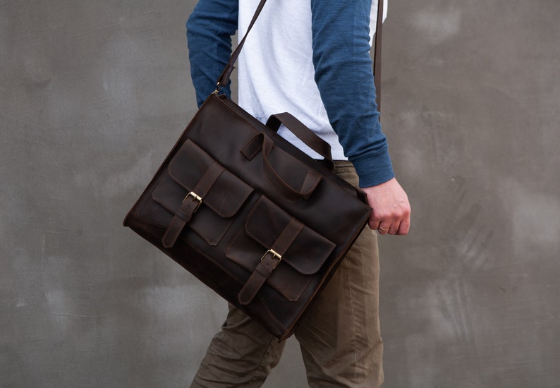 May include: A brown leather messenger bag with two front pockets and buckles. The bag has a long strap and is worn over the shoulder.