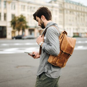 May include: A person wearing a gray shirt and brown leather backpack, holding a smartphone in their hand.