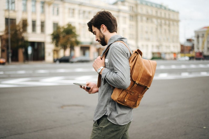 May include: A person wearing a gray shirt and brown leather backpack, holding a smartphone in their hand.