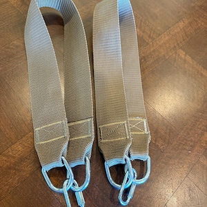 May include: Two tan-colored nylon straps with silver metal carabiner clips. The straps are looped at the top and have reinforced stitching at the bottom where the clips are attached. The background is a dark wood surface.