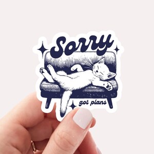 May include: A white cat is sleeping on a blue couch with the text "Sorry got plans" in a retro font. The sticker is 2.5 x 2.5 inches.
