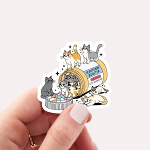 May include: A sticker of a pill bottle labeled "Serotonin Booster" with a bunch of cute cats spilling out of it. The bottle is yellow and blue with a red label. The cats are various colors and have different expressions. The sticker is 2.7 x 2.4 inches.