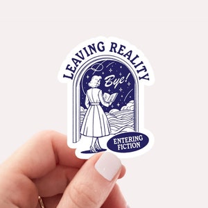 May include: A white sticker with a blue illustration of a woman reading a book, with the words "LEAVING REALITY" and "ENTERING FICTION". The sticker is 2.4 x 2.7 inches.