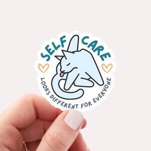 May include: A round sticker with a blue and white cat illustration. The cat is licking its paw. The text around the cat says "Self Care Looks Different For Everyone" with two small hearts on either side of the text.