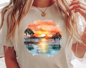 May include: White t-shirt with a watercolor sunset scene featuring palm trees and a body of water. The scene is surrounded by a light blue watercolor wash. The text "White" is visible in the bottom right corner of the image.
