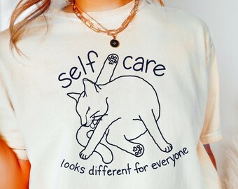 May include: A white t-shirt with a black line drawing of a cat grooming itself. The text "self care" is above the cat and the text "looks different for everyone" is below the cat.