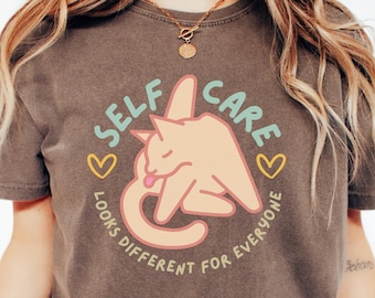 May include: A brown t-shirt with a graphic of a cat licking its paw and the text "Self Care Looks Different For Everyone".