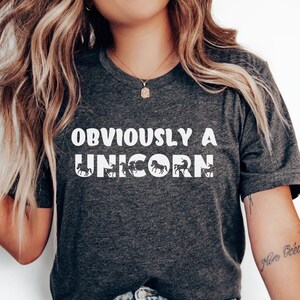 May include: A dark gray heather t-shirt with the text "OBVIOUSLY A UNICORN" in white. The shirt features a graphic of a unicorn with a rainbow mane.