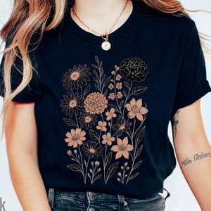 Op de afbeelding: Zwart t-shirt met een bloemenontwerp in perzik- en goudtinten. Het ontwerp bevat verschillende bloemen en bladeren. Het woord "Black" staat linksonder in de afbeelding.