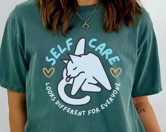 May include: Blue spruce t-shirt with a circular graphic that reads "SELF CARE LOOKS DIFFERENT FOR EVERYONE" and features a white cat licking itself. The shirt is a solid color and has short sleeves.
