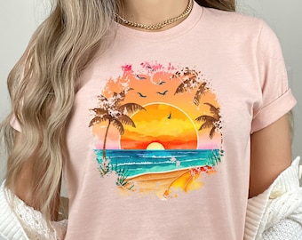 May include: A pink t-shirt with a graphic of a sunset over a beach with palm trees and birds flying overhead. The graphic is in a circle with a distressed, watercolor effect. The text "Heather Peach" is visible at the bottom of the image.