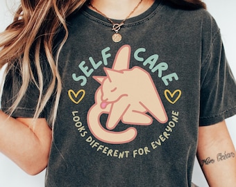 May include: A dark grey t-shirt featuring the text "SELF CARE" and "LOOKS DIFFERENT FOR EVERYONE" in a circular design. The graphic includes a cartoon cat in a relaxed pose, with yellow hearts. The shirt is a pepper colour.