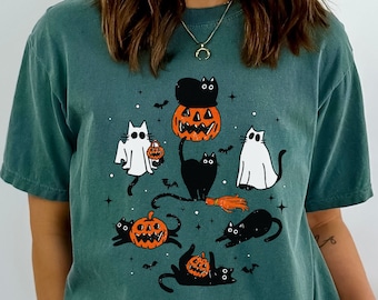 May include: A teal blue t-shirt with a graphic print of black cats and pumpkins. The graphic features a black cat sitting on top of a pumpkin with a smaller black cat on top of the cat. There are other black cats and pumpkins around the main graphic.