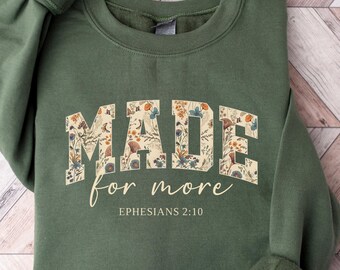 Könnte beinhalten: Militärgrünes Sweatshirt mit dem Schriftzug "MADE for more" in floralem Design. Der Text "Ephesians 2:10" steht darunter. Das Sweatshirt liegt gefaltet auf einer hellen Holzmaserung.
