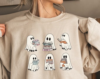 Puede incluir: Una sudadera beige con un estampado gráfico de seis fantasmas leyendo libros. Los fantasmas son blancos con ojos y bocas negros. Los libros son coloridos y están apilados. El texto "Sand" está impreso en la esquina inferior izquierda de la sudadera.