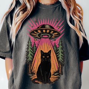 May include: Dark grey t-shirt featuring a black cat under a UFO with a pink and orange beam. The design includes green trees and the word "Pepper" at the bottom left. The shirt has short sleeves.