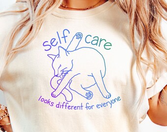 May include: A white t-shirt featuring a graphic of a cat grooming itself. The graphic is outlined in a rainbow gradient and the text "self care looks different for everyone" is below the cat.