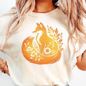 Może przedstawiać: Biały t-shirt z pomarańczową sylwetką lisa otoczonego kwiatami. Lis siedzi z ogonem owiniętym wokół ciała.