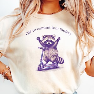 May include: A white t-shirt featuring a purple graphic of a raccoon riding a scooter. The text "Off to commit tom foolery" is printed in purple above the raccoon.