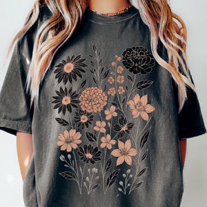 Peut inclure: T-shirt gris fonc&eacute; avec un motif floral en noir et p&ecirc;che. Le motif comprend diff&eacute;rents types de fleurs et de feuillage. Le mot "Pepper" est imprim&eacute; en bas &agrave; gauche du t-shirt.