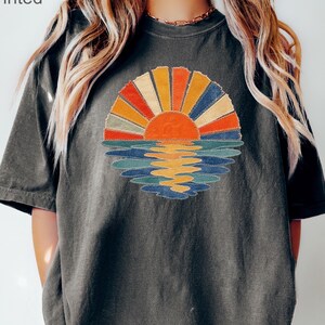 May include: A charcoal grey t-shirt featuring a graphic of a sun rising over water with rays in shades of orange, yellow, and blue. The design has a textured appearance with the text "not patch printed" and "Pepper" visible on the image.