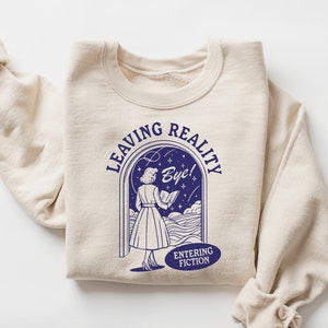 May include: A sand-colored sweatshirt with a blue graphic of a woman walking through a doorway with the text "Leaving Reality Bye! Entering Fiction".