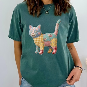 Op de afbeelding: Een blauwspar T-shirt met een patchwork kat ontwerp. De kat is gemaakt van verschillende stoffen met patronen, waaronder bloemen, ruitjes en ruitjes. De tekst "Blue Spruce" staat onderaan de afbeelding.