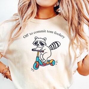 May include: A white t-shirt featuring a graphic of a raccoon riding a scooter. The text "Off to commit tom foolery" is printed above the graphic. The shirt is ivory in colour.